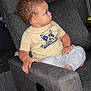 toddler, child, curly_hair, recliner, gray_furniture, indoor, brick_wall, wooden_floor, casual_clothing, tshirt, pants, barefoot, side_view, seated, home_interior, young_child, curious, looking_away, hand_on_armrest, cozy