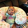 baby, smiling, onesie, floral_pillow, couch, blanket, infant, happy, child, indoor, cute, comfort, nursing_pillow, person, apparel, pattern, face, seated, home, soft