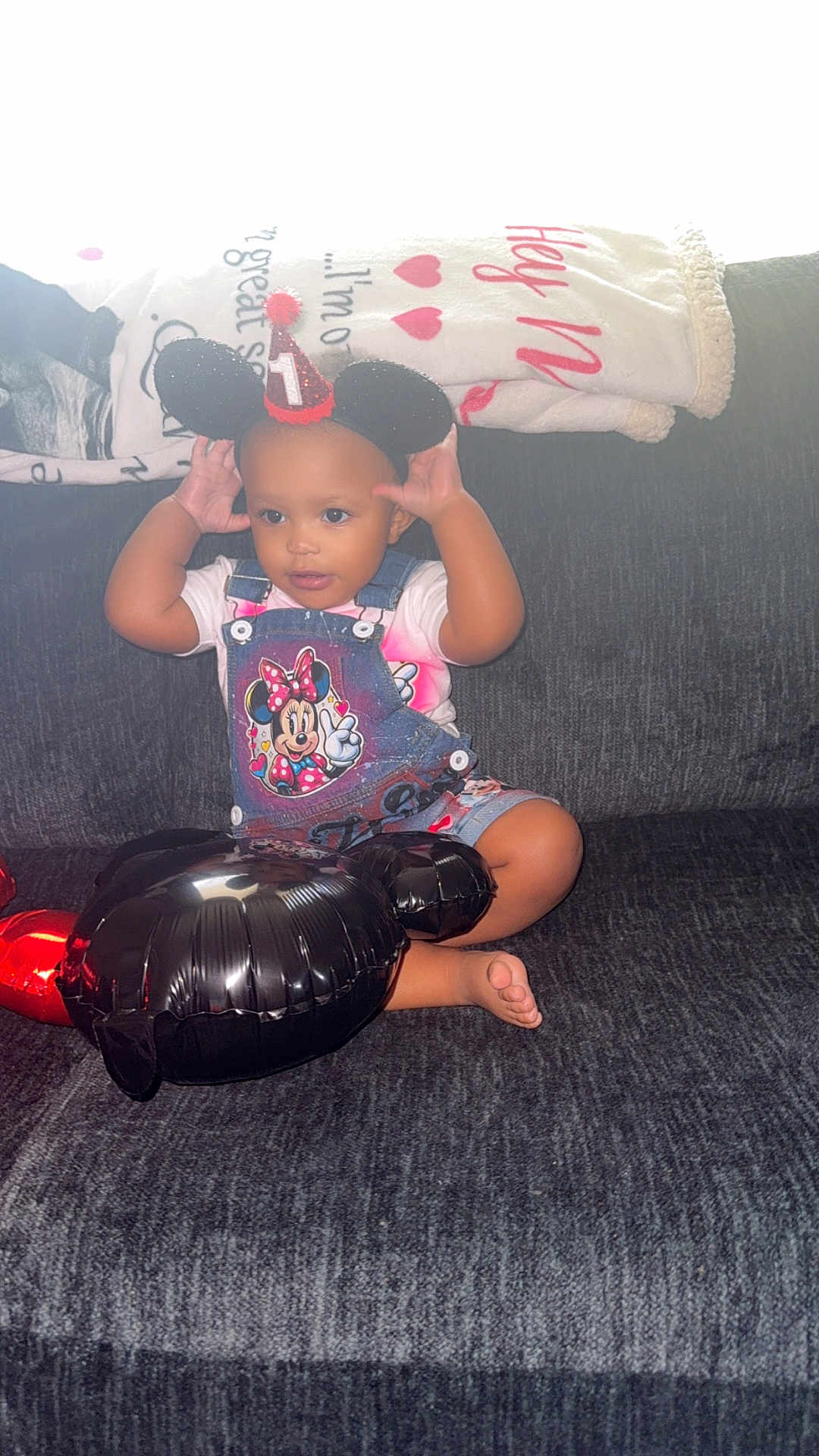 Mo Zyiah is registered to the contest to win money with this photo: toddler, child, party_hat, mouse_ears, balloon, minnie_mouse, denim_overalls, white_shirt, couch, blanket, indoor, cute, smiling, celebration, foot, black_balloon, red_accents, happy, young_child, casual_clothing