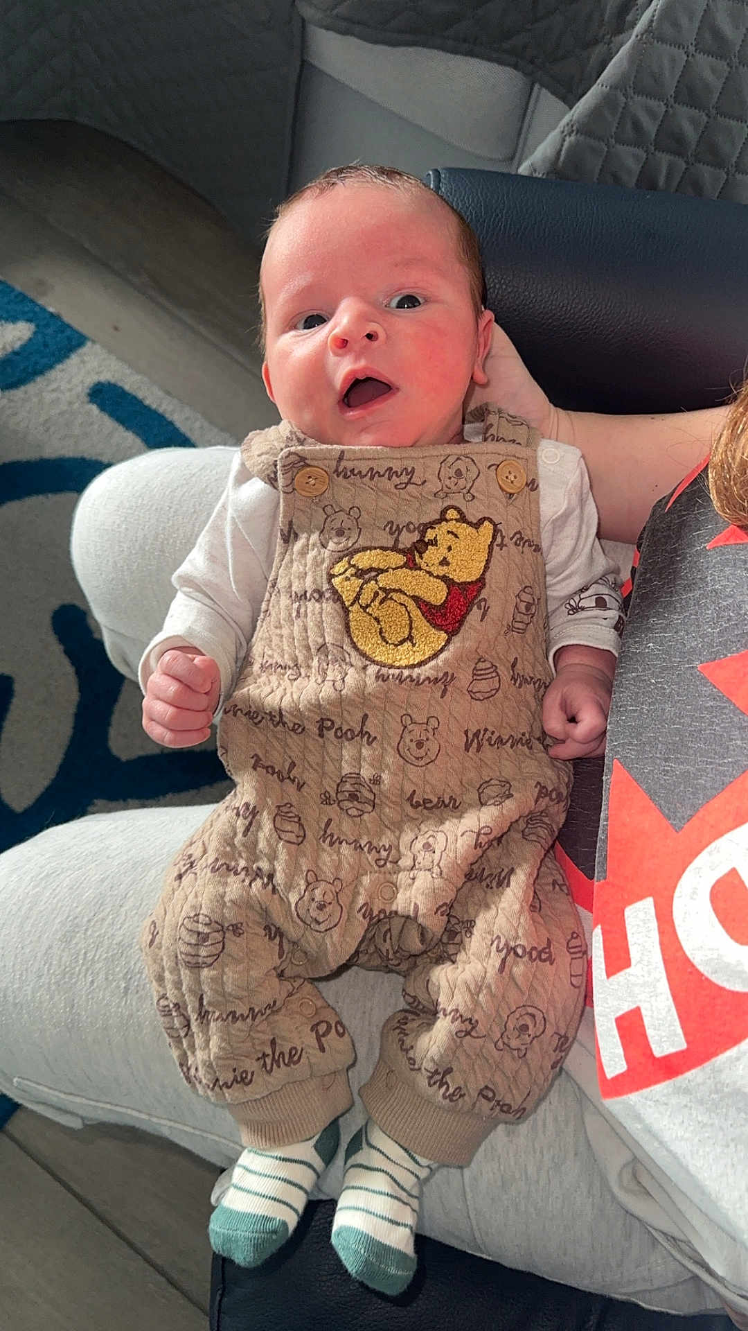 Devlin joined the competition — help win amazing prizes! baby, infant, winnie_the_pooh, overalls, face, open_mouth, wide_eyes, hand, arm, striped_socks, socks, couch, rug, floor, blanket, onesie, adult_partial, portrait, indoor, cute