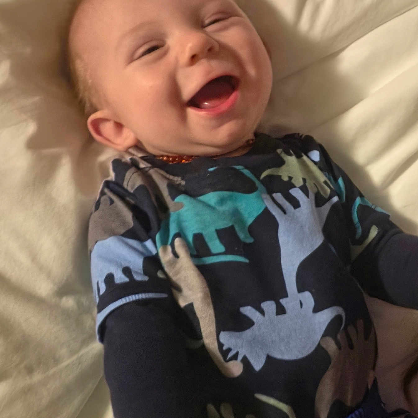 Levi is registered to the contest to win money with this photo: baby, infant, child, smiling, laughing, portrait, face, bed, bedding, indoors, cozy, pajamas, dinosaur_pattern, clothing, happy, lying_down, close_up, warm_lighting, brown_hair, home