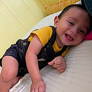Martin joined the competition — help win amazing prizes! baby, child, smiling, denim_overalls, yellow_shirt, bed, pillow, indoor, happy, cute, infant, person, skin, head, face, hand, leg, red_socks, cozy, portrait