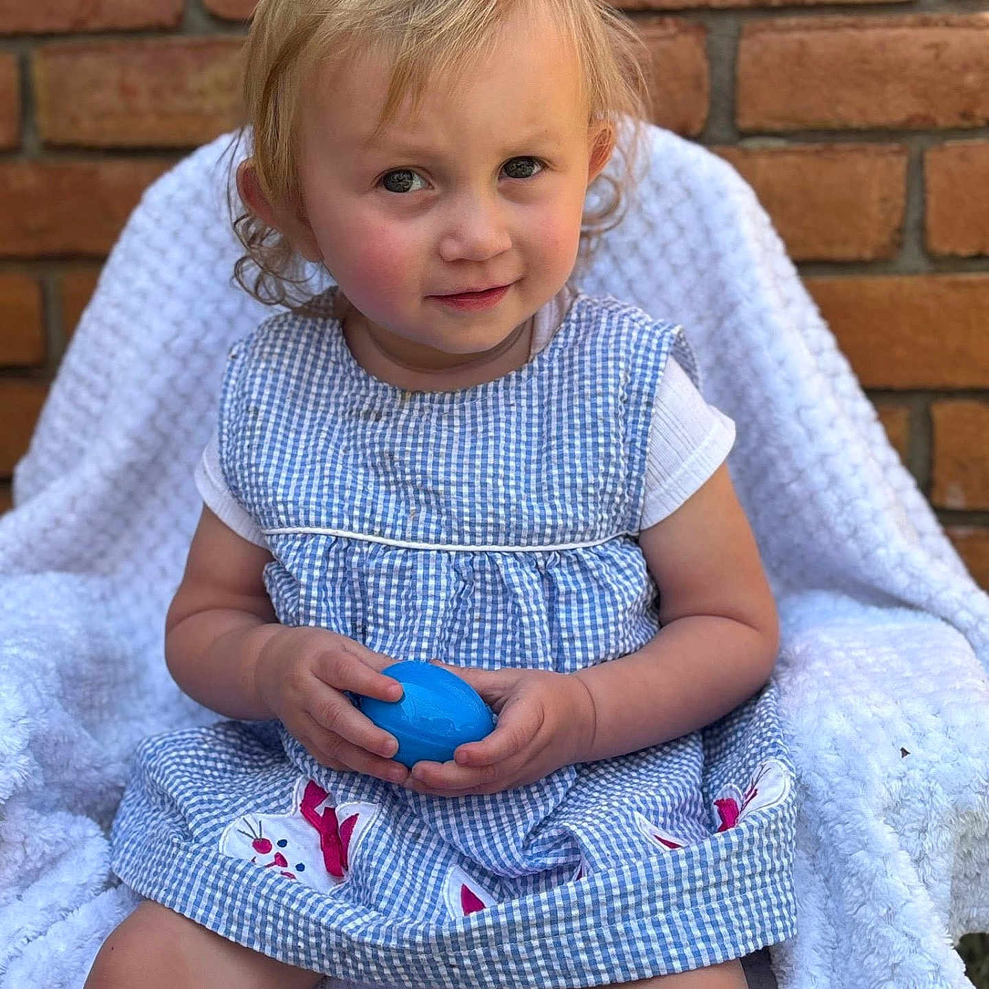 Isabella is registered to the contest to win money with this photo: blonde_hair, blue_egg, brick_wall, bunny_patches, chair, child, clothing, curly_hair, cute, gingham_dress, innocence, outdoor, person, playful, portrait, sitting, soft_lighting, toddler, white_blanket, young_child