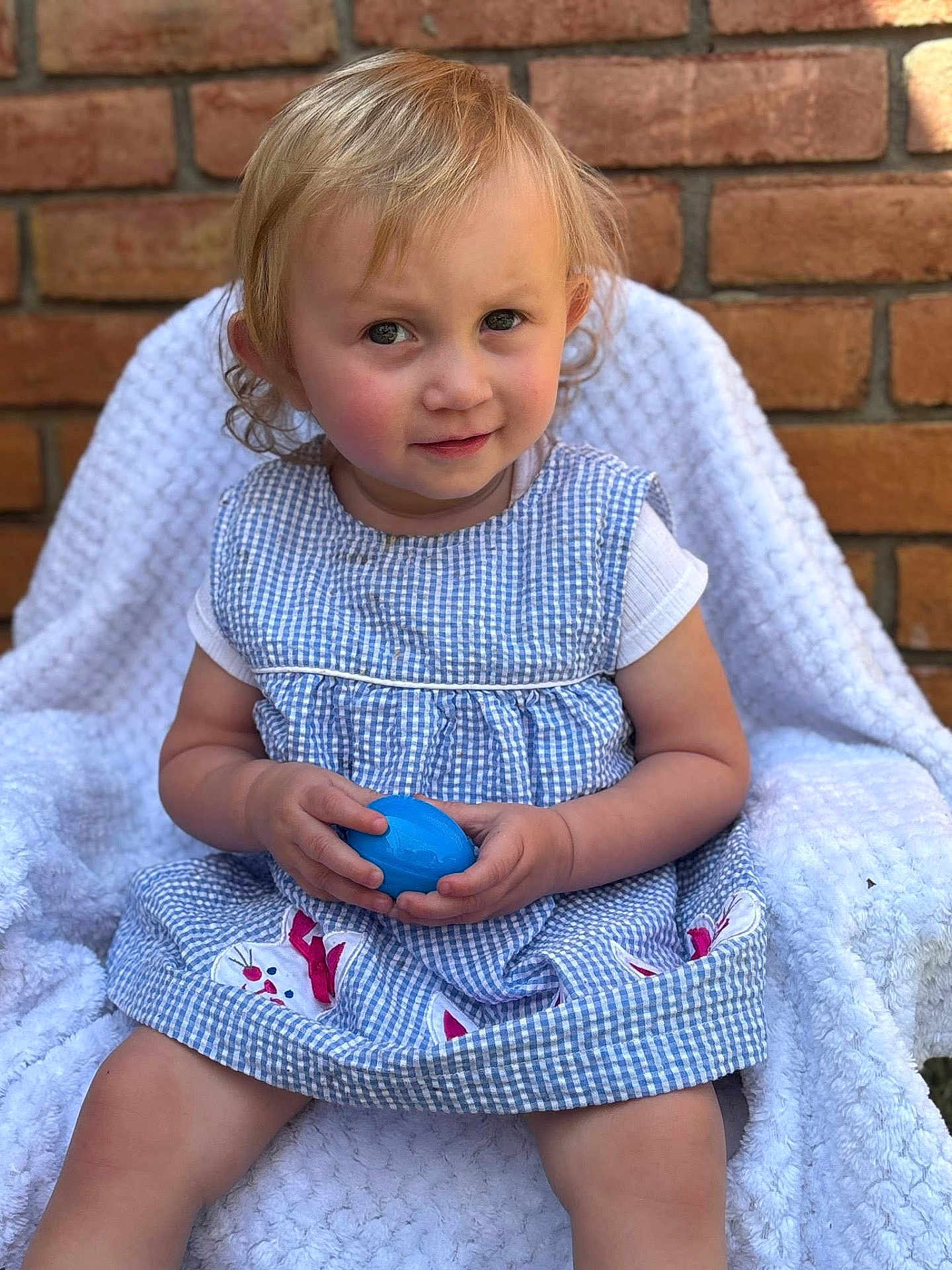 Isabella is registered to the contest to win money with this photo: toddler, child, blonde_hair, blue_egg, gingham_dress, bunny_patches, white_blanket, chair, brick_wall, outdoor, curly_hair, cute, portrait, sitting, playful, innocence, soft_lighting, clothing, person, young_child