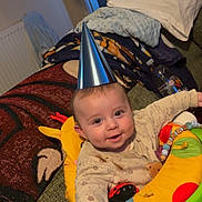 Enzo is registered to the contest to win money with this photo: baby, party_hat, smiling, indoor, activity_chair, blanket, toy, colorful, person, child, happy, face, clothing, hand, furniture, couch, living_room, head, infant, playtime