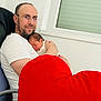 Mateo participe au concours pour gagner de l'argent avec cette photo : adult_male, newborn, baby, cuddling, red_heart_pillow, glasses, beard, white_t_shirt, arm, chair, window, indoor, sleeping, cozy, parent_child_interaction, minimalist_interior, soft_light, skin, portrait, affection
