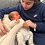 baby, newborn, sibling, girl, sleeping_baby, cuddly_toy, stuffed_animal, onesie, striped_pajamas, hoodie, nike_logo, watch, sofa, pillows, cozy, indoor, hand, family_moment, affection, portrait