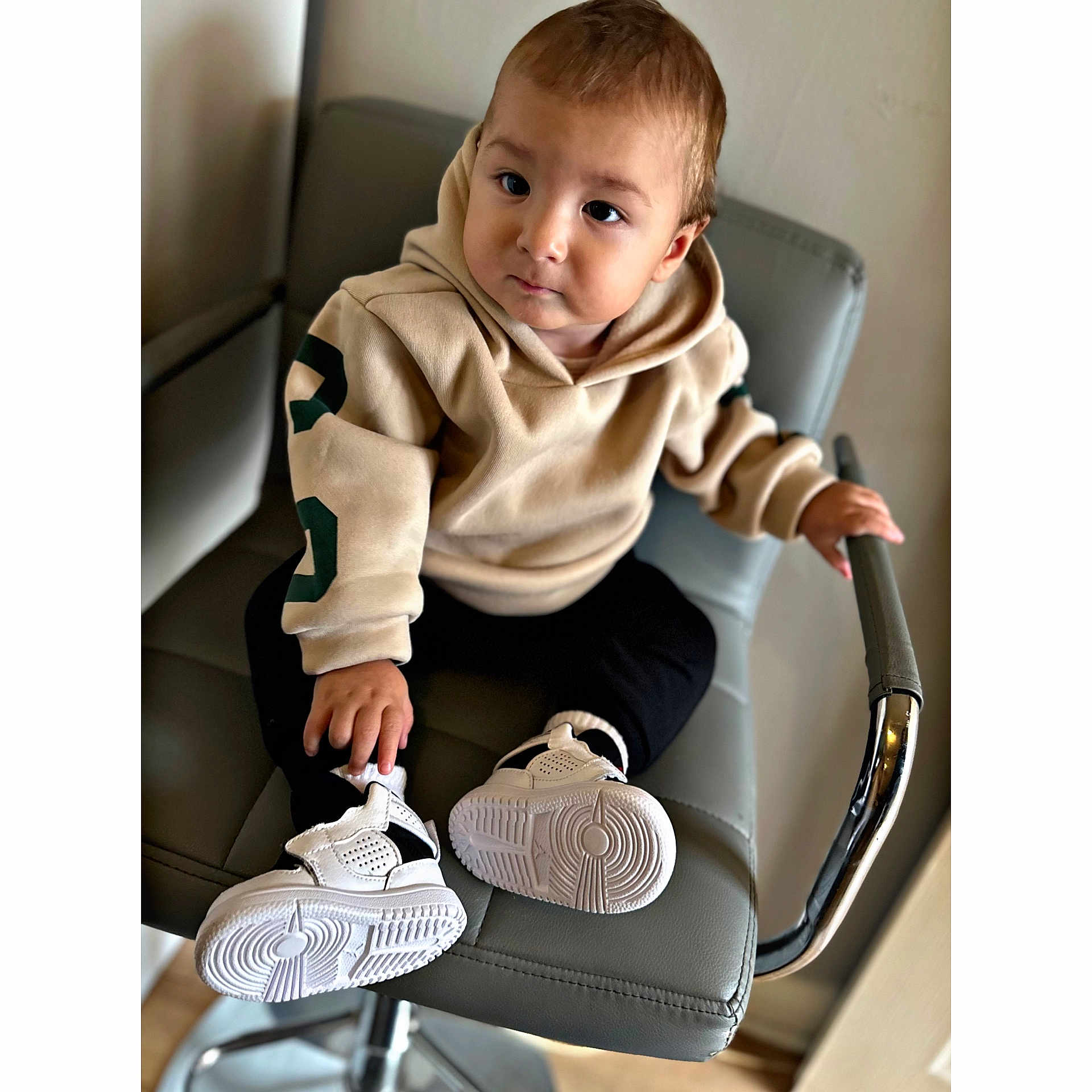 Renato is registered to the contest to win money with this photo: toddler, child, hoodie, sneakers, chair, indoor, person, footwear, clothing, seated, curious, modern_furniture, black_pants, white_shoes, hand, face, young_child, casual_wear, expression, light