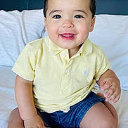 Levi is registered to the contest to win money with this photo: toddler, child, smiling, polo_shirt, yellow_shirt, blue_shorts, sitting, bed, white_bed, indoor, happy, cute, portrait, person, skin, face, hand, hair, shorts, clothing