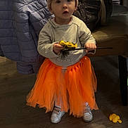 Thalya a rejoint le concours — aidez-le/la à gagner de superbes lots ! toddler, toy, orange_tutu, hat, indoor, wooden_floor, sweatshirt, child, person, table, jacket, shoes, curious, boy, clothing, furniture, play, toy_car, wand, floor