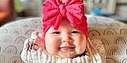 Isabella is registered to the contest to win money with this photo: apparel, baby, cheeks, child, clothing, cushion, cute, face, hand, happy, headband, home, indoors, infant, portrait, rainbow_pattern, red_bow, sitting, smiling, soft_lighting