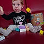 Aria a rejoint le concours — aidez-le/la à gagner de superbes lots ! toddler, child, blue_eyes, curly_hair, black_shirt, text_on_clothing, building_blocks, red_couch, socks, playing, indoor, toy, cardboard_box, person, face, legs, seated, motion_blur, home, casual