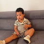 casual_wear, child, curly_hair, gray_couch, holding_object, home_interior, indoor, legs_spread, looking_away, person, polo_shirt, portrait, relaxed, shorts, sitting, soft_lighting, striped_clothing, toddler, white_shoes, young_child