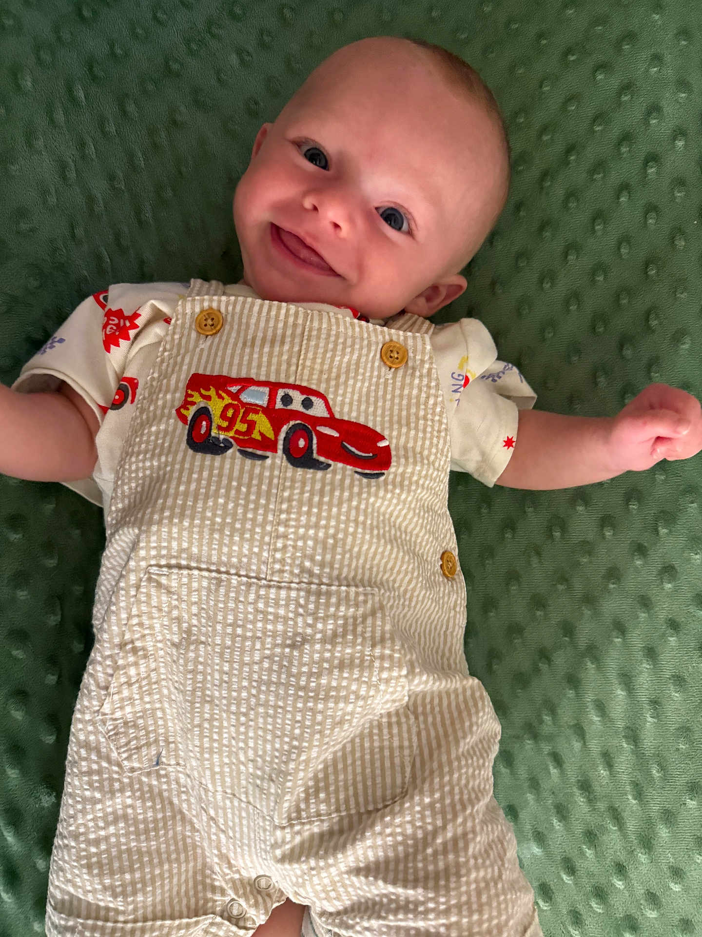 Grayson joined the competition — help win amazing prizes! baby, smiling, overalls, cartoon_car, green_blanket, cute, infant, clothing, happy, face, child, playful, texture, fabric, button, tongue_out, portrait, lying_down, casual, young