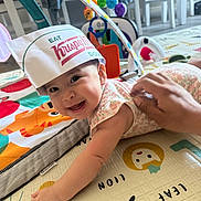 Augustina is registered to the contest to win money with this photo: baby, infant, smiling, hat, paper_hat, play_mat, toy, hand, home, floor, colorful, cute, child, person, indoor, happy, reaching, clothing, support, face