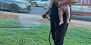 Augustina is registered to the contest to win money with this photo: elderly_person, baby, woman, child, yard, grass, plants, hose, watering, suburban, fence, tree, car, sidewalk, house, outdoor, daylight, person, holding, footwear
