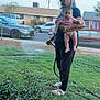 elderly_person, baby, woman, child, yard, grass, plants, hose, watering, suburban, fence, tree, car, sidewalk, house, outdoor, daylight, person, holding, footwear