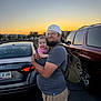 man, baby, child, glasses, cap, backpack, parking_lot, car, sunset, smile, casual_clothing, outdoor, facial_hair, holding, happy, person, vehicle, road, evening, urban