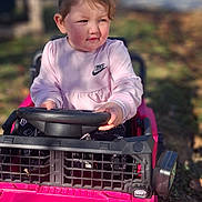 Melissa is registered to the contest to win money with this photo: toddler, child, toy_car, pink, outdoor, play, pigtails, steering_wheel, sunlight, casual_clothing, nike_logo, grass, daylight, expression, person, young_child, vehicle, playtime, fun, nature