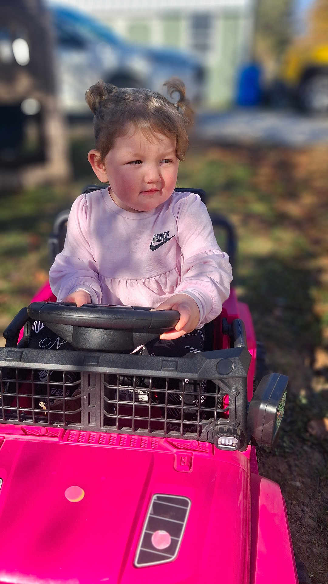 Melissa is registered to the contest to win money with this photo: toddler, child, toy_car, pink, outdoor, play, pigtails, steering_wheel, sunlight, casual_clothing, nike_logo, grass, daylight, expression, person, young_child, vehicle, playtime, fun, nature