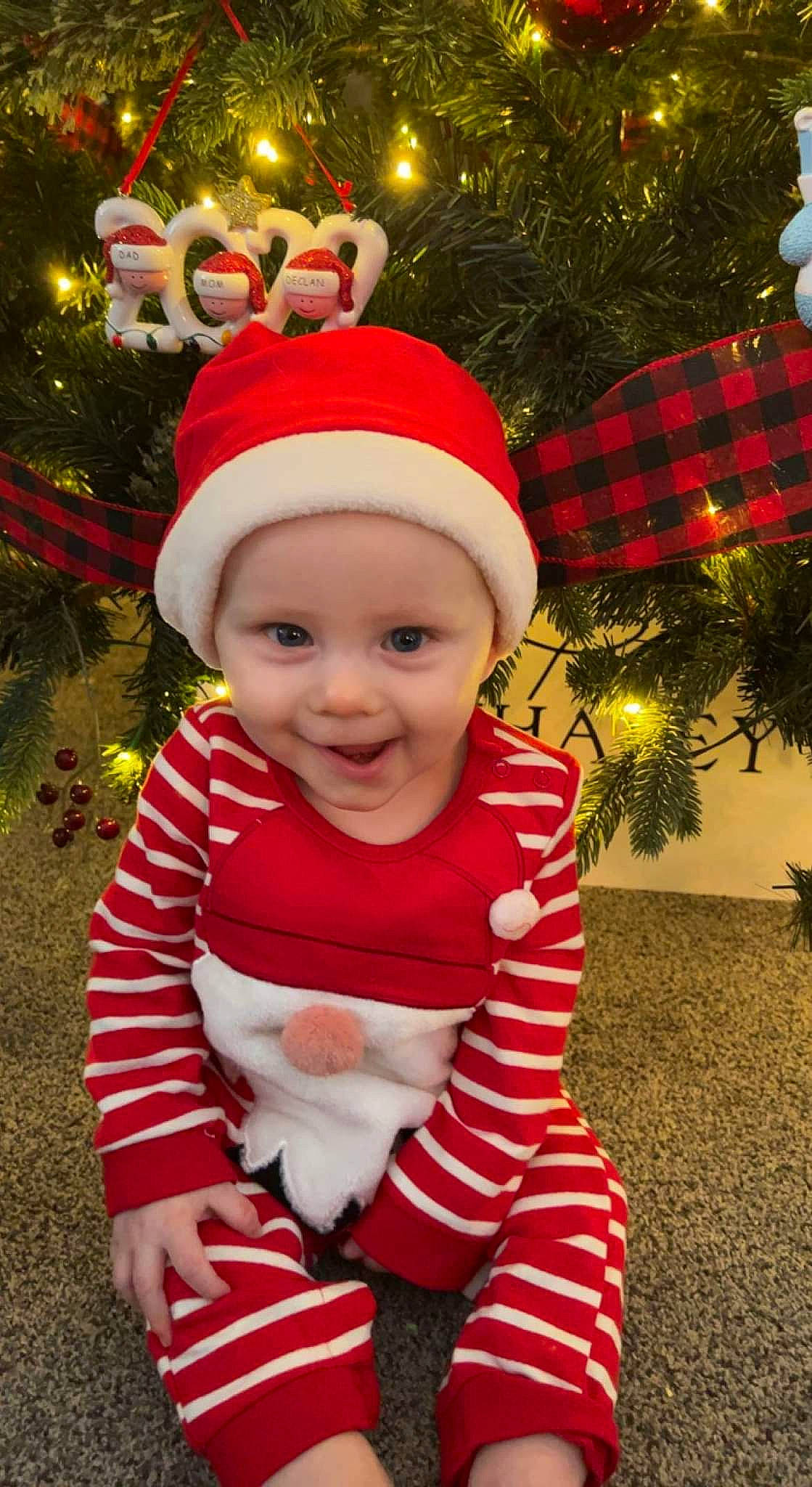 Declan is registered to the contest to win money with this photo: baby_toddler_clothing, cap, christmas, christmas_decoration, christmas_ornament, christmas_tree, event, face, fun, happy, headwear, holiday, holiday_ornament, human_body, joy, light, ornament, person, red, sleeve