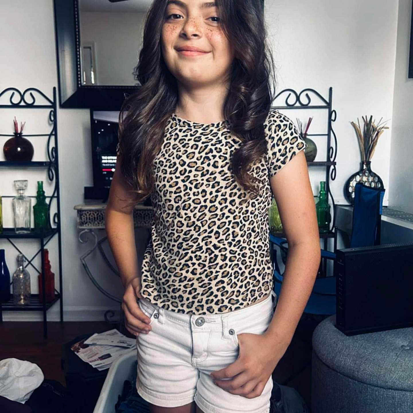 Keithlyn Strother joined the competition — help win amazing prizes! bottles, casual_clothing, ceiling_fan, child, curly_hair, decor, freckles, furniture, girl, home, indoor, leopard_print, living_room, mirror, person, posing, shelves, smile, standing, white_shorts
