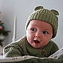 baby, child, knit_hat, green_sweater, blanket, face, wide_eyes, open_mouth, cute, indoor, christmas_tree, holiday, soft_texture, warm_clothing, portrait, infant, cozy, expression, decor, festive
