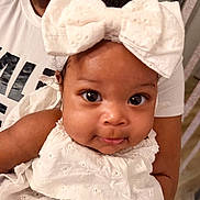 Anyla is registered to the contest to win money with this photo: baby, child, infant, white_bow, white_dress, curly_hair, wide_eyes, smile, person, indoor, holding, adult, face, portrait, closeup, cute, clothing, headwear, skin, expression