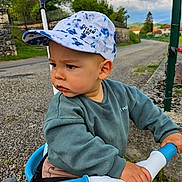 Pharell participe au concours pour gagner de l'argent avec cette photo : toddler, child, hat, cap, tricycle, handlebars, outdoor, road, grip, greenery, clouds, sky, casual_clothing, curious, young_child, daylight, nature, expression, person, transportation