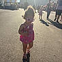 child, toddler, sunglasses, curly_hair, pink_clothing, sandal, bottle, shadow, outdoor, sunlight, people, fairground, tent, asphalt, summer, daytime, walking, family, casual, crowd