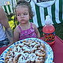 Celine joined the competition — help win amazing prizes! child, curly_hair, pink_dress, funnel_cake, powdered_sugar, fruit_juice, beverage_bottle, table, outdoor, festival, green_banner, white_stripes, face, skeptical_expression, tattooed_arm, summer, food, snack, grass, daylight