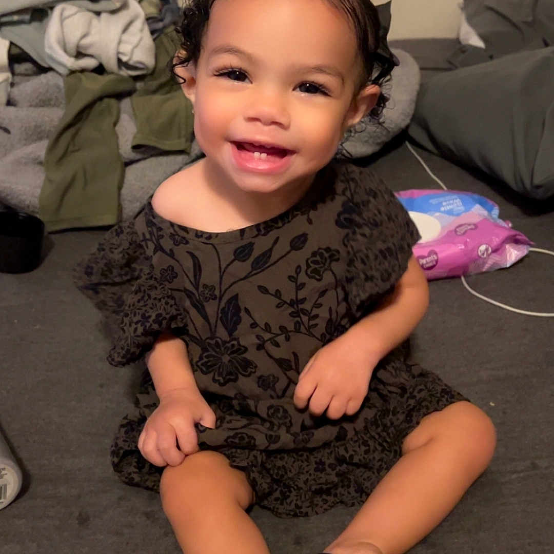 Kiari Bennett joined the competition — help win amazing prizes! black_hair, black_shoes, blanket, cheerful, child, clothing, couch, curly_hair, cute, dress, face, happy, home, indoor, person, sitting, small_hands, smiling, toddler, young_child