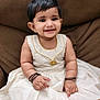 baby, child, smiling, dress, white_dress, golden_border, bracelets, necklace, sitting, couch, brown_couch, indoors, portrait, happy, cute, person, black_hair, infant, jewelry, skin