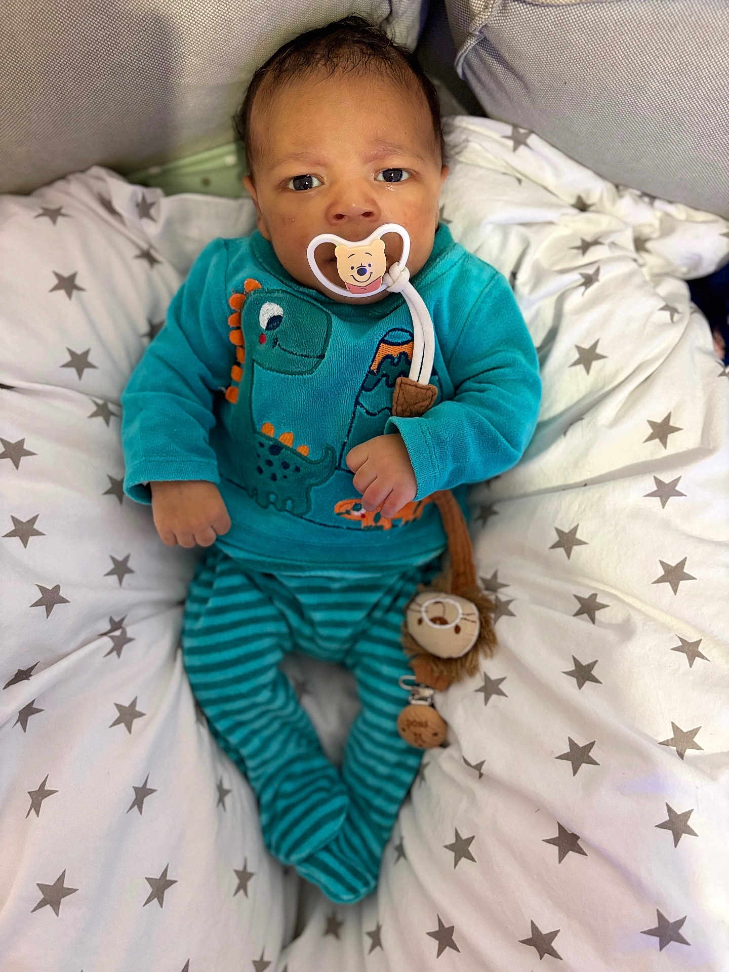 Fodé a rejoint le concours — aidez-le/la à gagner de superbes lots ! baby, infant, pacifier, plush_toy, dinosaur_outfit, striped_pants, star_pattern, cushion, blanket, blue_clothing, cozy, cute, indoor, child, person, lying_down, comfortable, soft_toys, face, young