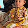 toddler, child, onesie, colorful_blanket, indoor, television, furniture, smiling, cute, face, sitting, sock, plush, patterned_clothing, living_room, toy, cozy, home, yellow, playful
