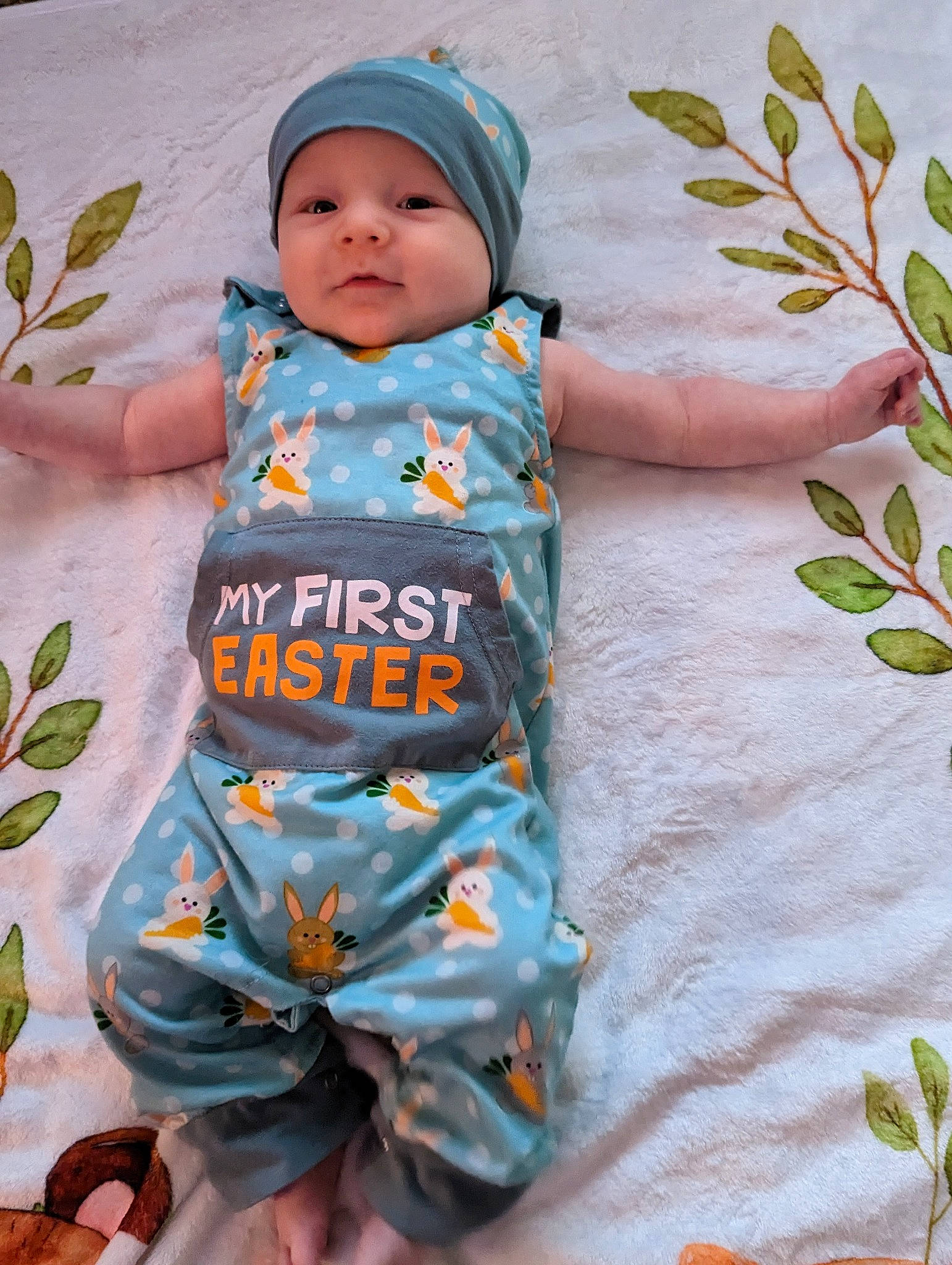 Flynn is registered to the contest to win money with this photo: baby, baby_products, baby_toddler_clothing, cap, child, doll, face, grass, happy, headwear, linens, outerwear, pattern, peach, people_in_nature, person, plant, sitting, skin, sleeve