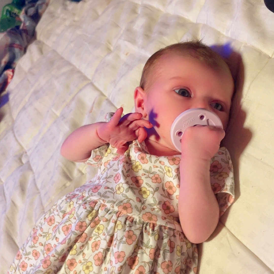 Haylo is registered to the contest to win money with this photo: baby, bed, blanket, blue_eyes, child, cute, face, floral_dress, hand, indoors, infant, light_shadow, lying_down, newborn, pacifier, person, quilt, resting, sleepwear, soft_light