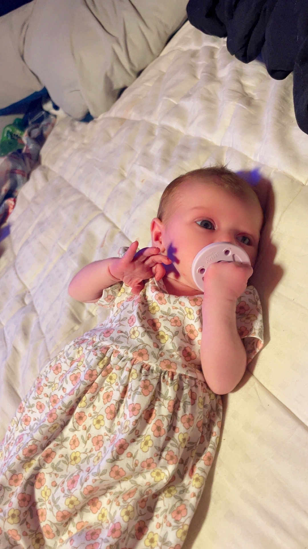 Haylo is registered to the contest to win money with this photo: baby, infant, pacifier, bed, quilt, floral_dress, hand, face, blue_eyes, light_shadow, blanket, sleepwear, person, child, indoors, resting, soft_light, cute, newborn, lying_down