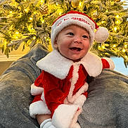 Ezjahramos is registered to the contest to win money with this photo: baby, christmas_tree, christmas_lights, christmas_hat, red_clothing, white_trim, blanket, smiling, holiday, festive, indoor, cute, sitting, happy, toddler, soft_texture, decorations, cozy, portrait, seasonal