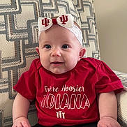 Josephine is registered to the contest to win money with this photo: baby, child, infant, red_shirt, headband, bow, smile, indoor, chair, patterned_fabric, cute, person, young_child, happy, sitting, portrait, clothing, face, hand, eyes