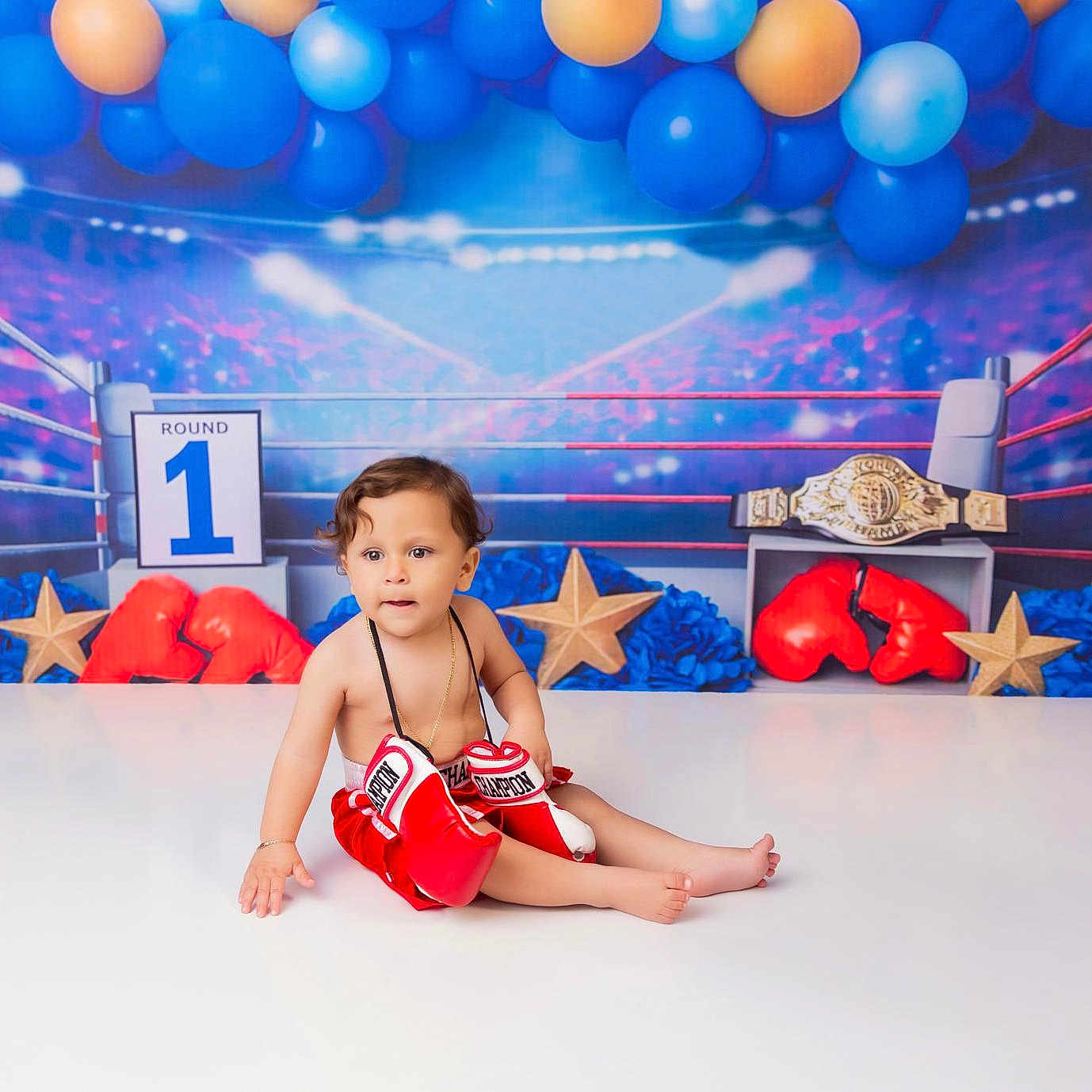 Christopher is registered to the contest to win money with this photo: baby, child, boxing_gloves, red_shorts, sitting, balloons, blue_balloons, orange_balloons, boxing_ring, championship_belt, stars, decorations, round_1_sign, indoor, portrait, cute, playful, floor, person, toddler
