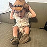 toddler, child, cowboy_hat, hat, sitting, couch, indoor, curious, casual_clothing, shoes, brown, beige, portrait, young_child, person, furniture, home, cute, small_child, expression