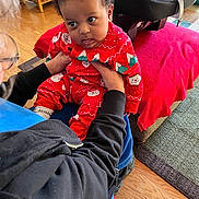 Tatum is registered to the contest to win money with this photo: baby, child, infant, pajamas, red_clothing, adult_hands, living_room, car_seat, blanket, floor, wood_floor, person, face, curly_hair, seated, indoor, carpet, furniture, holding, black_jacket