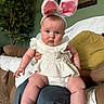baby, child, bunny_ears, white_clothing, sitting, indoor, living_room, plant, green_wall, framed_picture, person, couch, carpet, cute, portrait, blue_eyes, plush, hands, legs, soft_lighting