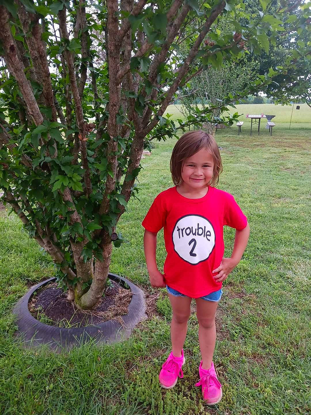 Olivia is registered to the contest to win money with this photo: botany, evergreen, garden, grass, groundcover, happy, joy, leaf, magenta, people_in_nature, person, plant, recreation, shorts, shrub, smile, t_shirt, terrestrial_plant, toddler, tree