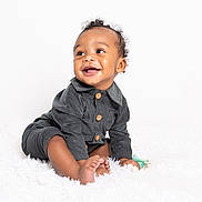 Khionyx joined the competition — help win amazing prizes! baby, infant, smiling, happy, sitting, dark_grey_outfit, pacifier, toy, curly_hair, white_background, fluffy_rug, cute, child, portrait, studio_photo, skin, clothing, foot, hand, expression