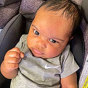 Nehemiah is registered to the contest to win money with this photo: baby, infant, car_seat, grey_clothing, onesie, fist, face, eyes, head, person, child, seat_belt, fabric, pattern, closeup, indoor, expression, skin, hair, casual_wear