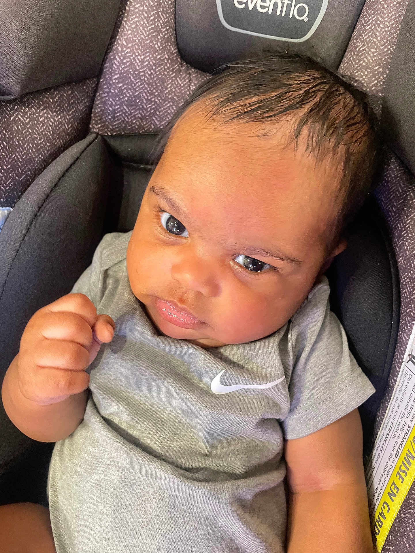 Nehemiah is registered to the contest to win money with this photo: baby, infant, car_seat, grey_clothing, onesie, fist, face, eyes, head, person, child, seat_belt, fabric, pattern, closeup, indoor, expression, skin, hair, casual_wear
