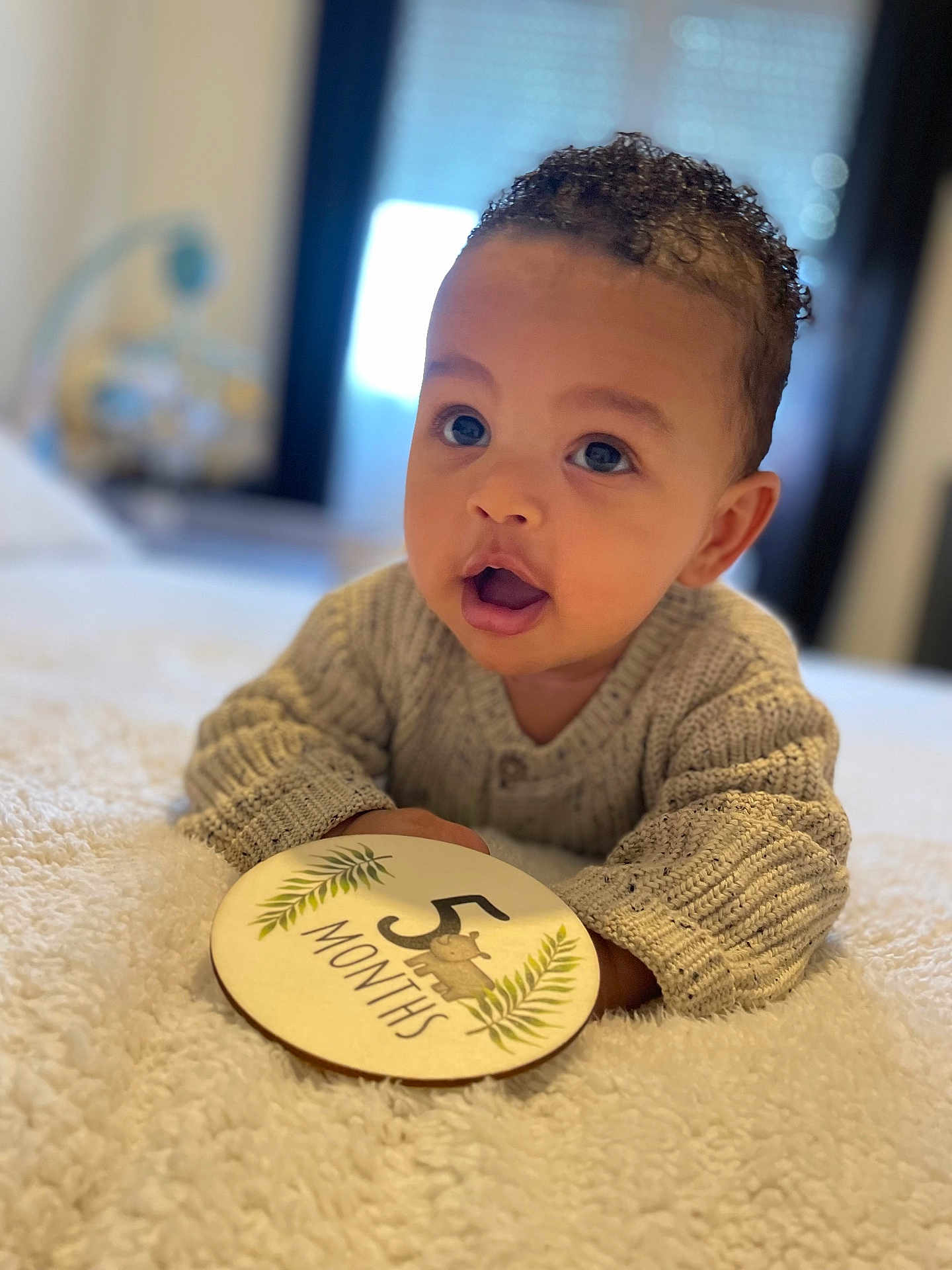 Sloann a rejoint le concours — aidez-le/la à gagner de superbes lots ! baby, infant, 5_months, milestone, curly_hair, big_eyes, sweater, blanket, cute, portrait, smiling, lying_down, indoor, soft_focus, child, person, cozy, warm, hands, closeup