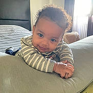 Sloann a rejoint le concours — aidez-le/la à gagner de superbes lots ! baby, child, curly_hair, striped_clothing, pillow, bedroom, window, sunlight, smile, hands, cozy, indoor, headboard, blanket, furniture, infant, person, portrait, cute, relaxing